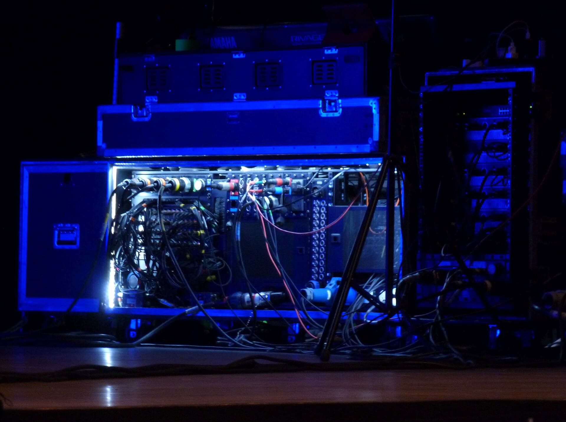 Rear of a concert mixing console and amplifier racks lit blue.